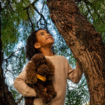 A young girl with curly dark hair stands in a tree trunk, looking upward with a smile while holding a brown curly-haired plush dog wearing a yellow collar. She is wearing a beige knit sweater and black pants, set against a background of green leaves and tree bark.