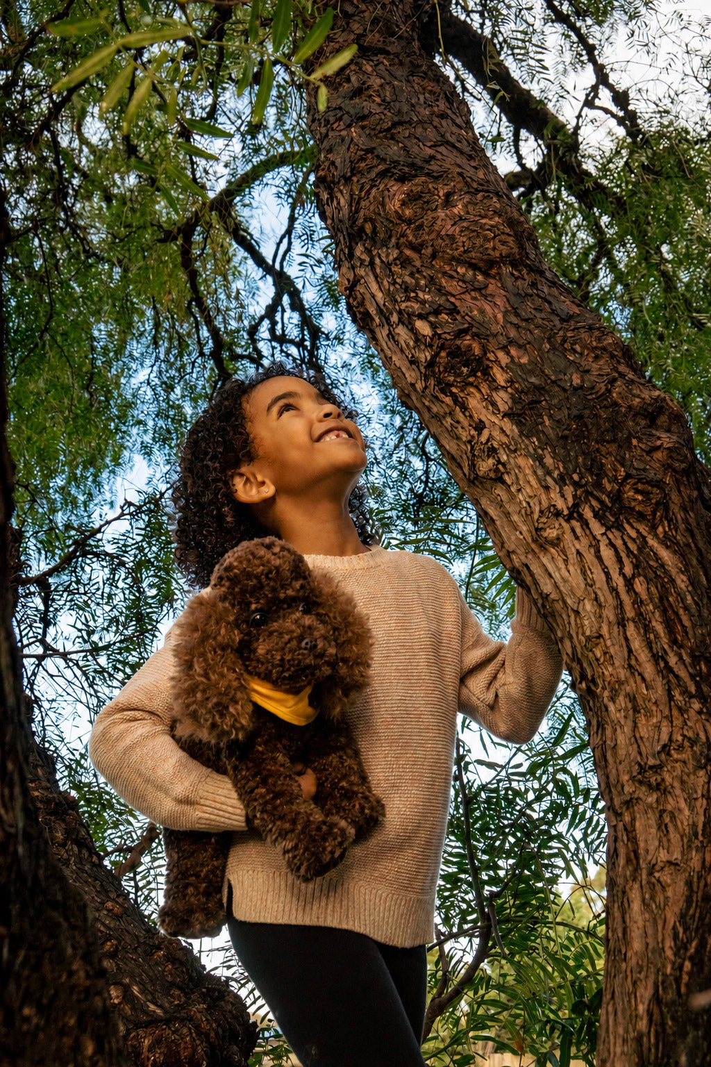 A young girl with curly dark hair stands in a tree trunk, looking upward with a smile while holding a brown curly-haired plush dog wearing a yellow collar. She is wearing a beige knit sweater and black pants, set against a background of green leaves and tree bark.
