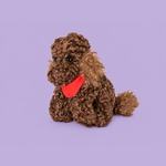 A brown curly-haired plush dog wearing a bright red bandana, sitting against a solid light purple background.