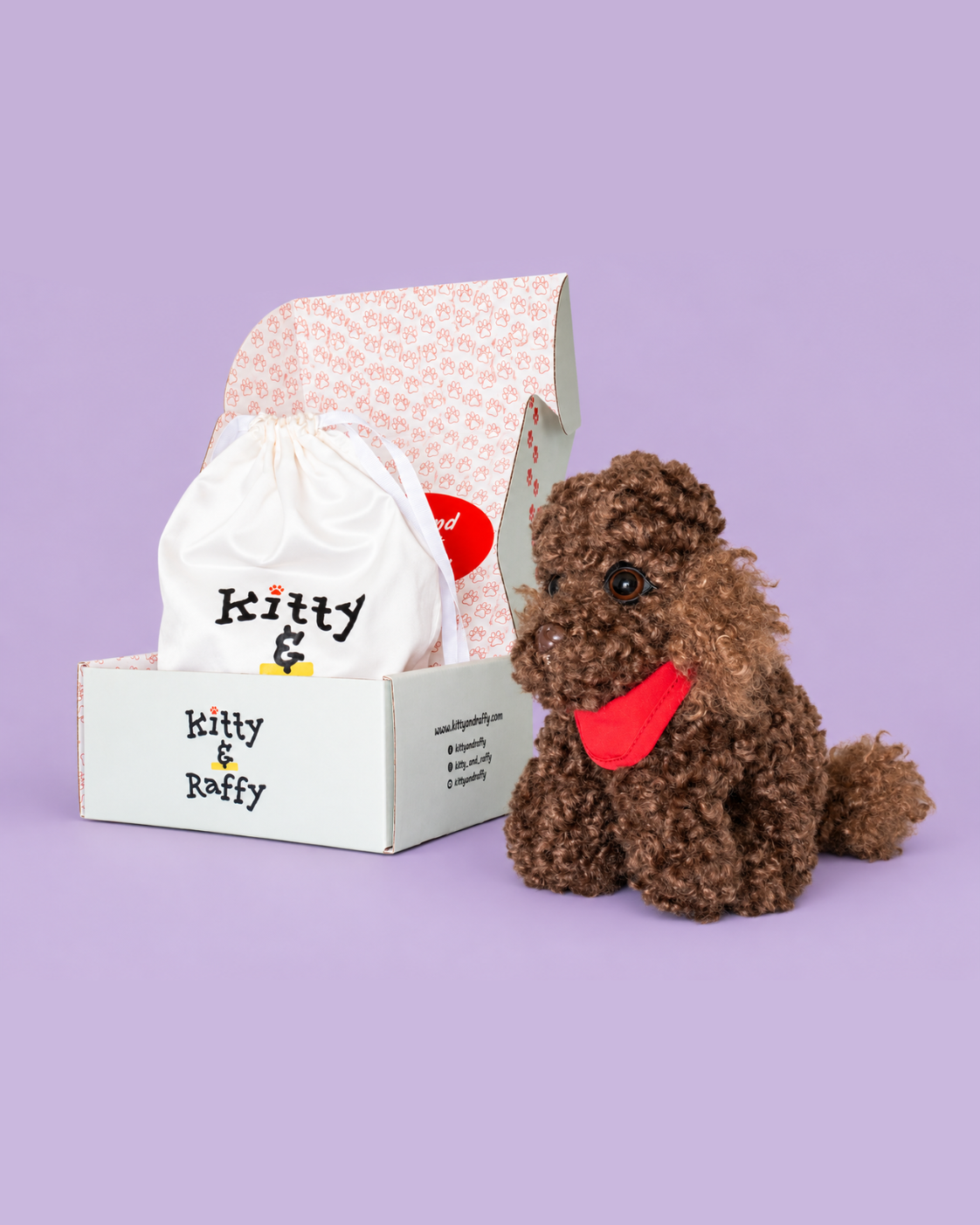A brown curly-haired plush dog toy wearing a red bandana sits next to its branded white box and drawstring bag on a solid lavender background.