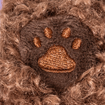 Close-up of a brown furry plush dog with an embroidered brown paw print patch in the center, set against a light purple background.
