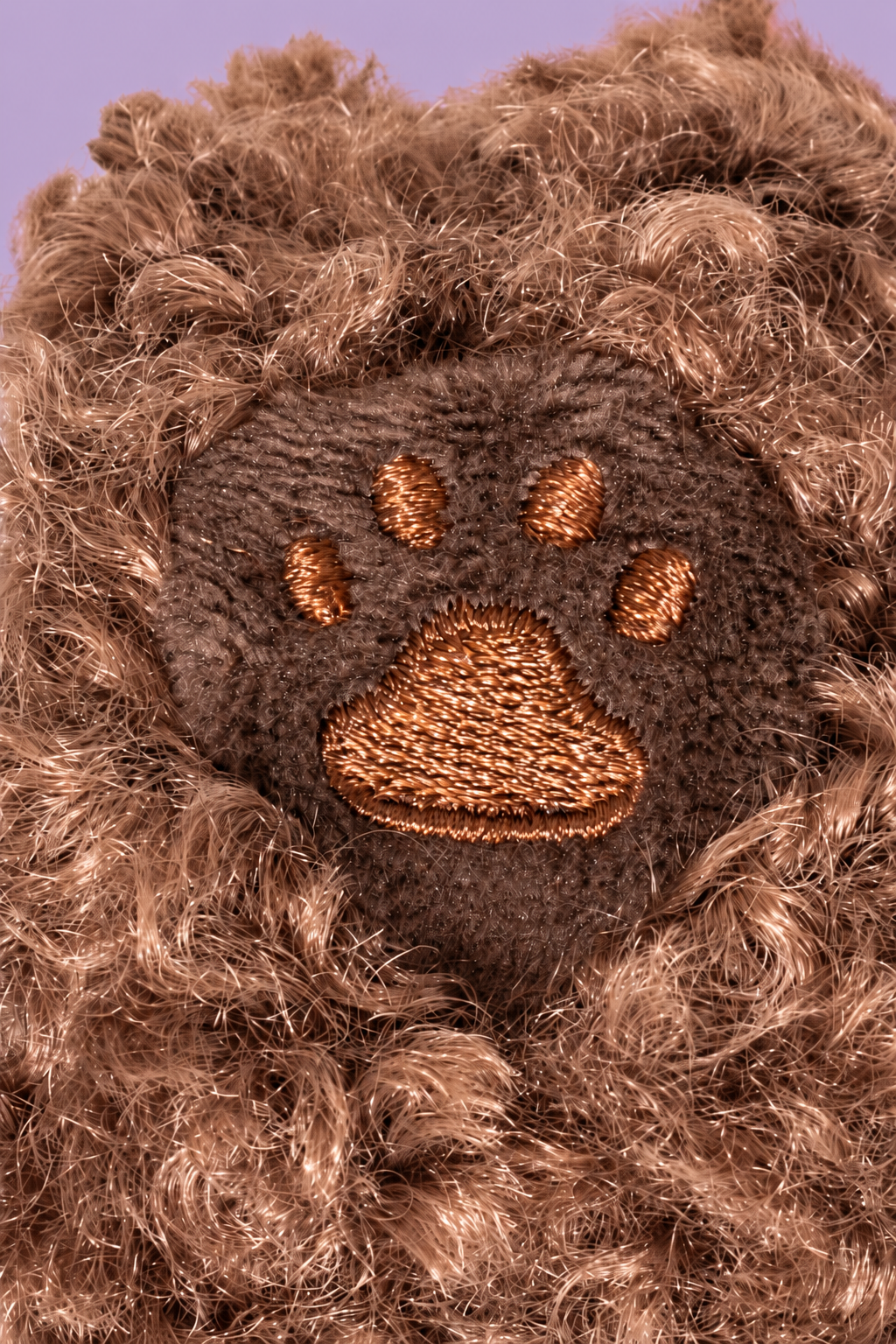 Close-up of a brown furry plush dog with an embroidered brown paw print patch in the center, set against a light purple background.