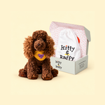 A brown curly-haired plush dog toy sits beside an open branded gift box containing a white drawstring bag printed with 'Kitty & Raffy' on a warm cream background.